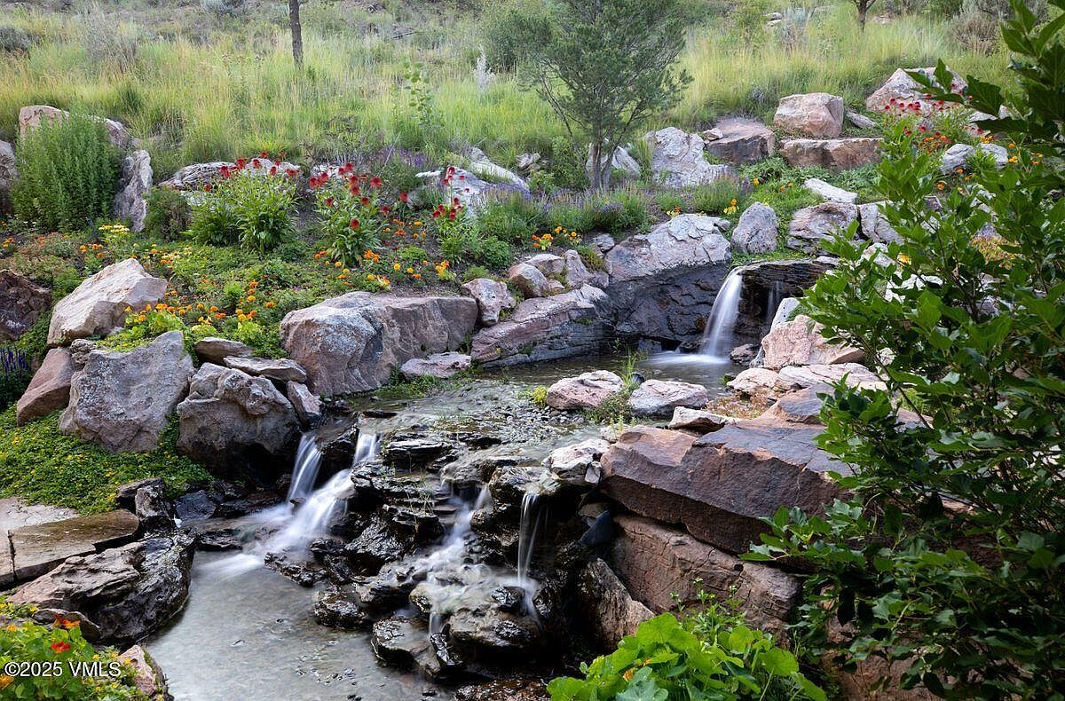 This image showcases a beautifully landscaped yard featuring a multi-tiered rock garden with cascading waterfalls. Lush greenery and colorful flowers accent the natural stone, creating a serene and inviting outdoor space. The water feature adds a dynamic element, enhancing the property's appeal.