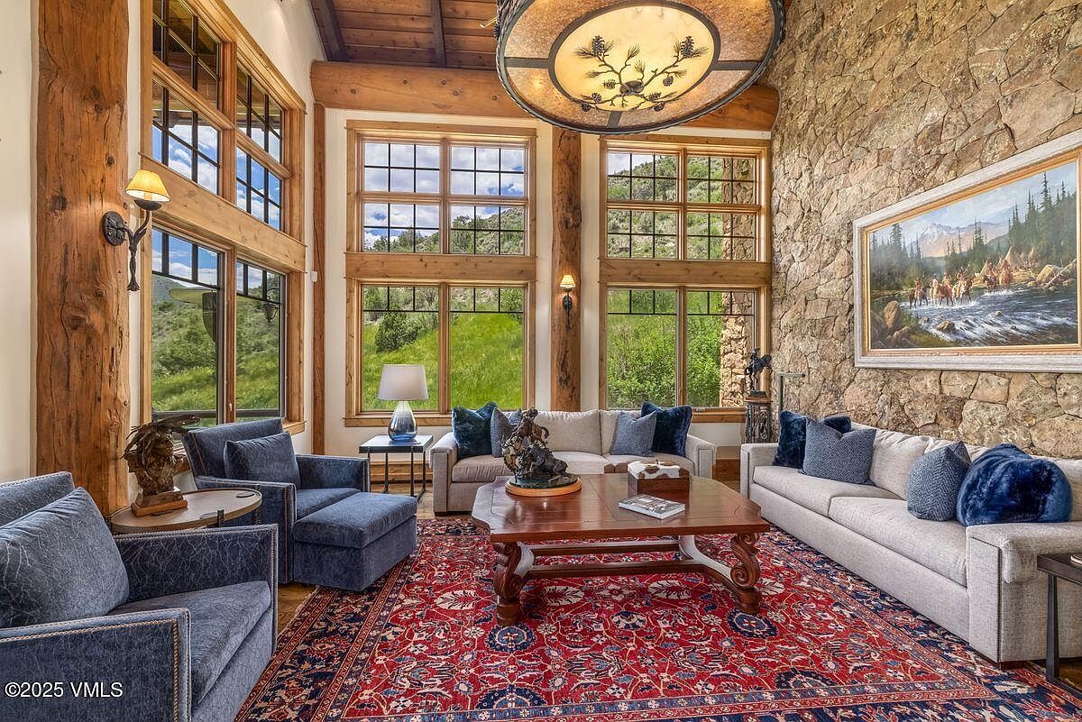 This is a spacious living room featuring a stone accent wall, large windows with natural light, and comfortable seating arrangements. The room is decorated with a mix of textures and patterns, including a large oriental rug, creating a warm and inviting atmosphere. The wooden beams and chandelier add to the rustic charm of the space.