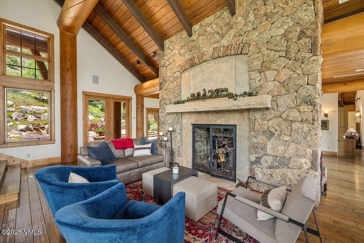 This is an inviting living room featuring a stone fireplace as a focal point, complemented by exposed wooden beams on the ceiling. The room is furnished with comfortable seating, including blue armchairs and a sofa, arranged around a central coffee table and ottoman. Natural light streams in through the windows, enhancing the warm and cozy atmosphere of the space.