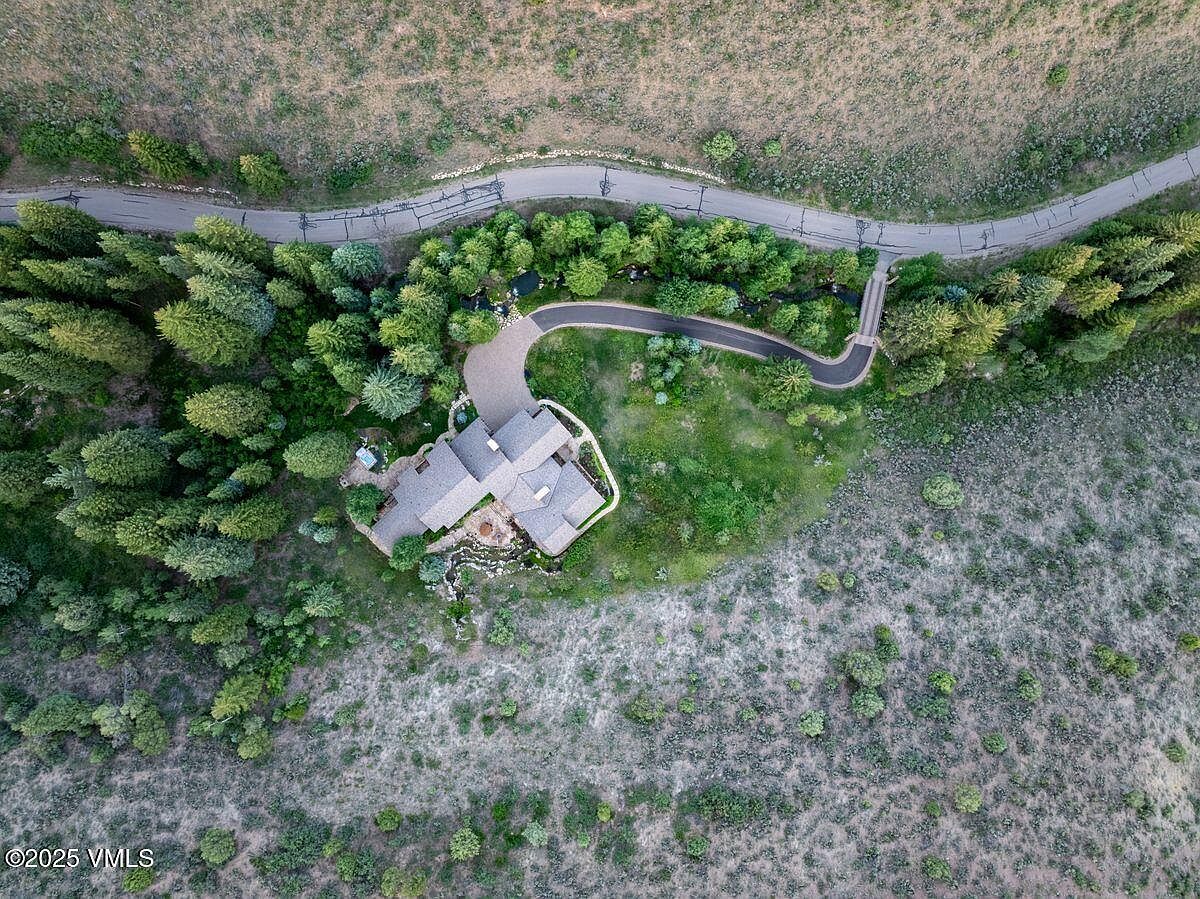 This aerial shot showcases a luxurious home nestled amidst lush greenery and natural terrain. A winding driveway leads to the property, which features a multi-gabled roof and a stone-accented exterior. The surrounding landscape includes mature trees, manicured lawns, and natural vegetation, creating a sense of privacy and tranquility.