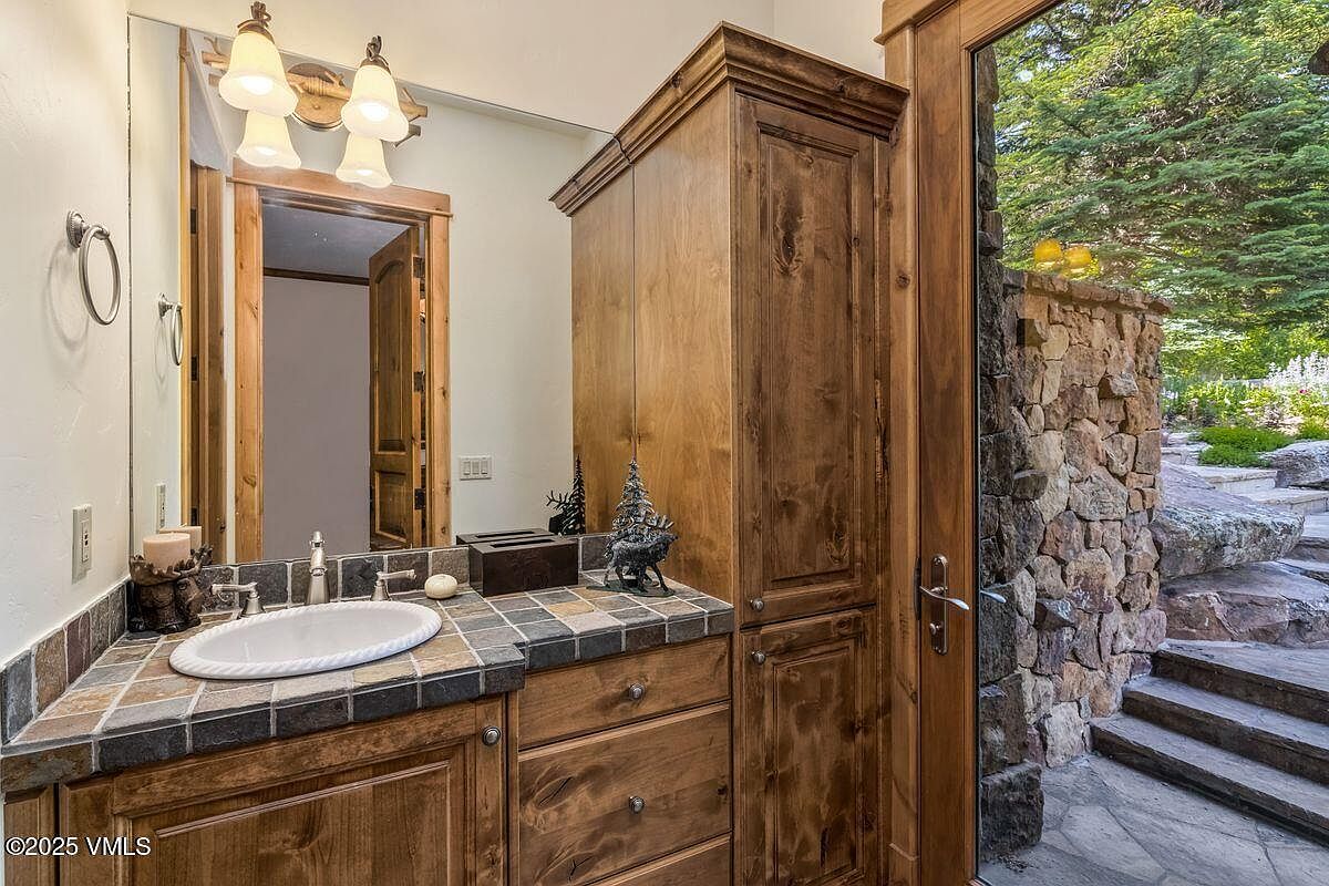 This is a well-appointed guest bathroom featuring a wooden vanity with a tiled countertop and an oval sink. A large mirror reflects the vanity and a glimpse into another room. The bathroom has a rustic yet elegant feel, with a door leading to an outdoor area with stone steps and greenery.