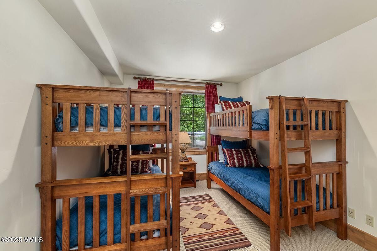 This is a bedroom featuring two wooden bunk beds with blue bedding and decorative pillows. A small nightstand with a lamp sits between the beds, and a patterned rug lies on the carpeted floor. The room has a cozy and rustic feel, suitable for a children's room or guest bedroom in a mountain home.
