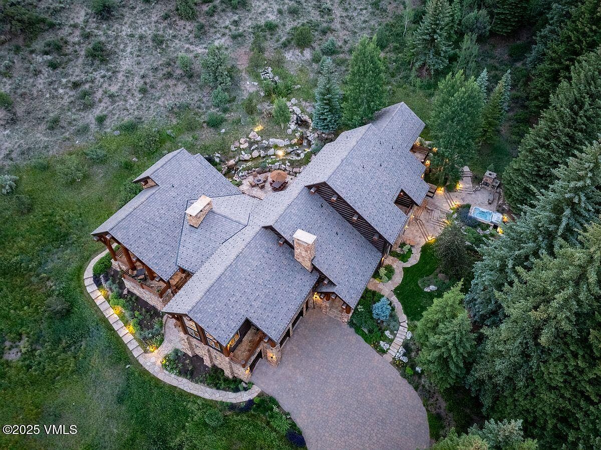This aerial view showcases a luxurious mountain home with a stone and timber exterior, complemented by a meticulously landscaped yard. The property features a winding driveway, multiple rooflines, stone chimneys, and outdoor living spaces including a patio with a hot tub and a water feature. The surrounding landscape includes mature trees and natural vegetation, enhancing the property's privacy and appeal.