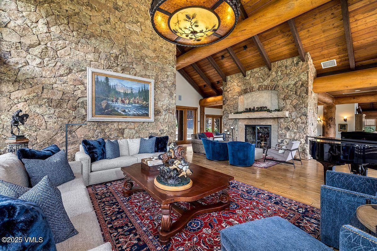 This is an interior shot of a luxurious living room featuring a stone wall and fireplace, complemented by exposed wooden beams on the ceiling. The room is furnished with comfortable sofas adorned with blue pillows, a wooden coffee table, and a grand piano, creating a warm and inviting atmosphere. A large area rug adds a touch of elegance to the hardwood floors.