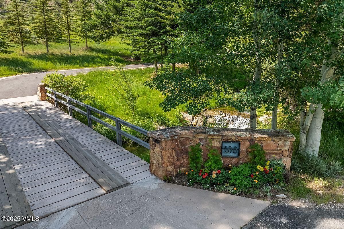This image showcases a charming yard/garden area with a wooden bridge leading to a stone structure displaying the address '100'. Lush greenery, including trees and flowering plants, surrounds the area, creating a serene and inviting atmosphere. A small waterfall can be seen in the background, adding to the natural beauty of the property.