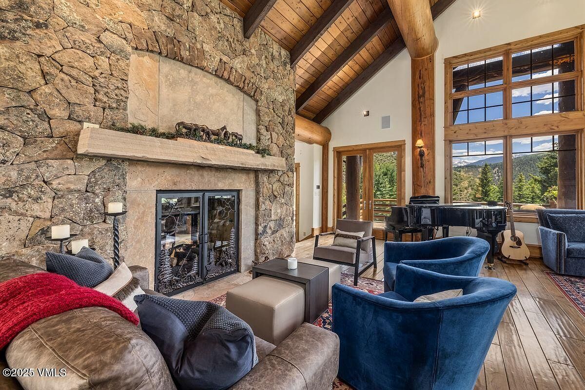 This is an interior shot of a luxurious living room featuring a stone fireplace with a mantel adorned with decorative horses. The room has exposed wooden beams on the ceiling, large windows offering a view of the outdoors, and comfortable seating including plush blue armchairs and a leather sofa. A grand piano sits near the windows, adding to the room's elegance.