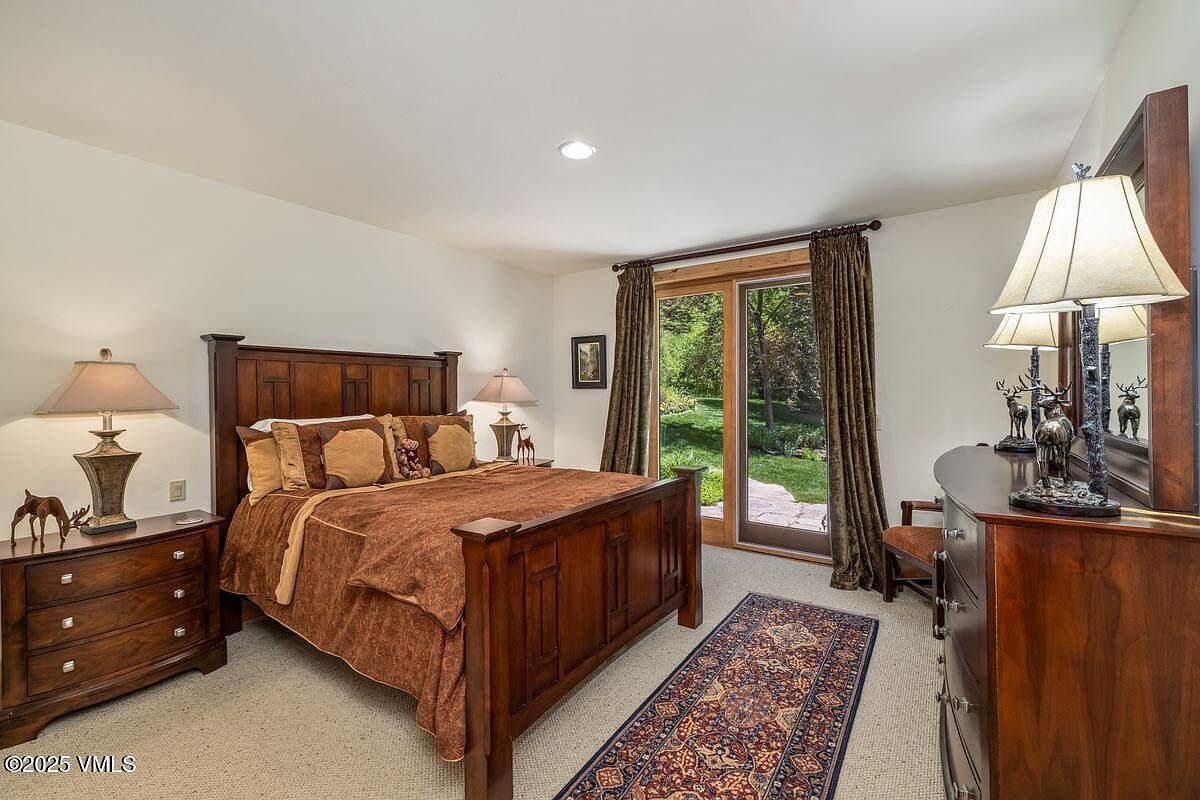 This is a warm and inviting guest bedroom featuring a wooden bed frame with a brown patterned comforter and decorative pillows. The room includes matching wooden nightstands and a dresser, complemented by lamps and decorative items. A sliding glass door with curtains provides natural light and a view of the outdoors, enhancing the room's cozy and comfortable atmosphere.