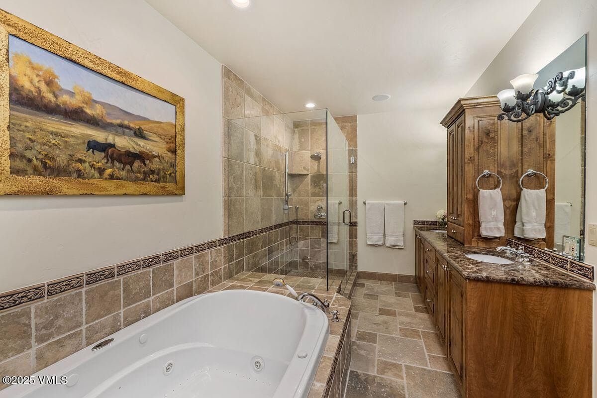 This is a well-appointed primary bathroom featuring a soaking tub, a glass-enclosed shower, and a vanity with a marble countertop. The cabinetry is a rich wood tone, and the walls are adorned with a landscape painting. The overall impression is luxurious and relaxing.