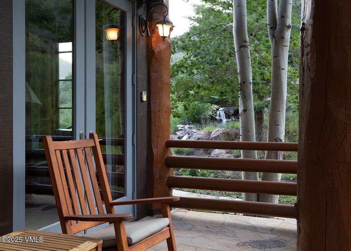 This image showcases a charming patio or balcony area, featuring a wooden rocking chair with a cushioned seat, suggesting a relaxing outdoor space. The patio is constructed with stone tiles, and a wooden railing provides a rustic touch. Through the railing, a view of trees and a small waterfall can be seen, enhancing the natural ambiance.