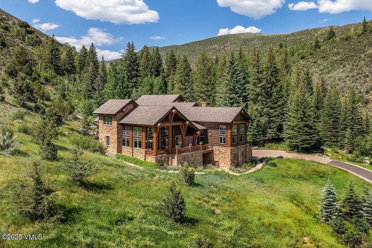 This image showcases the front exterior of a luxurious mountain home. The house features a combination of stone and wood construction, with a prominent covered porch supported by large wooden beams. The surrounding landscape includes lush greenery, mature trees, and a winding driveway, creating a sense of privacy and natural beauty.