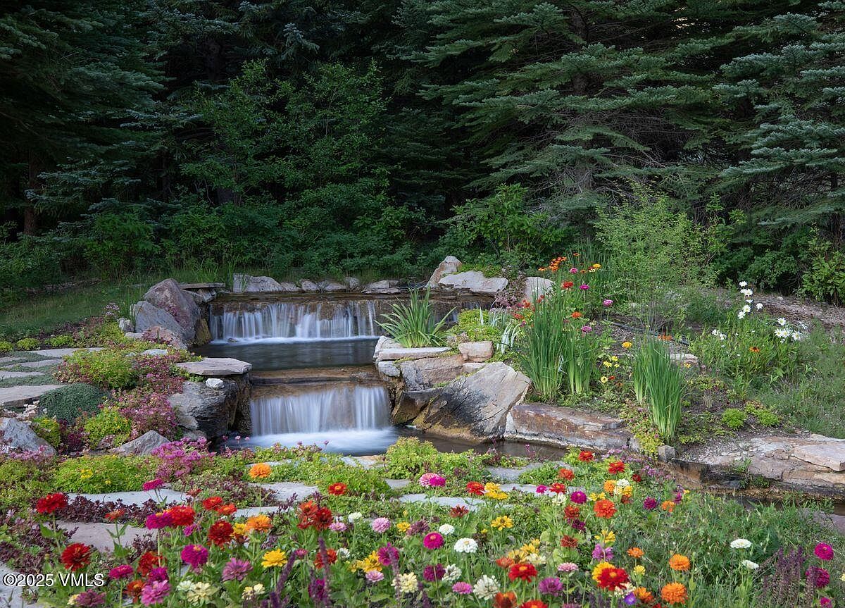 This image showcases a beautifully landscaped yard featuring a multi-tiered waterfall cascading over natural rocks. Vibrant, colorful flowers bloom in the foreground, adding a lively touch to the scene. The backdrop consists of lush green trees, creating a serene and inviting outdoor space.