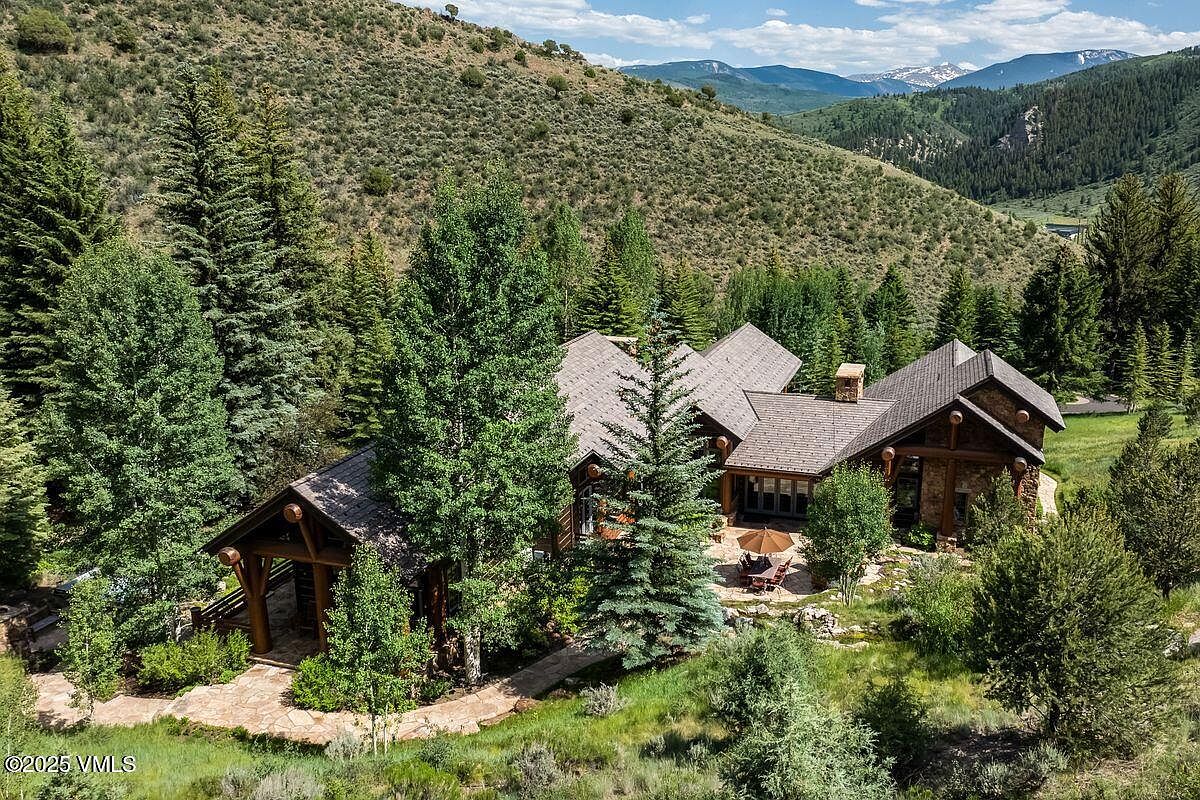 This aerial view showcases a luxurious mountain home nestled among lush greenery and towering trees. The architecture features a blend of natural stone and wood, with a complex roofline and inviting outdoor patio area. The surrounding landscape includes rolling hills and distant mountain peaks, creating a serene and private setting.