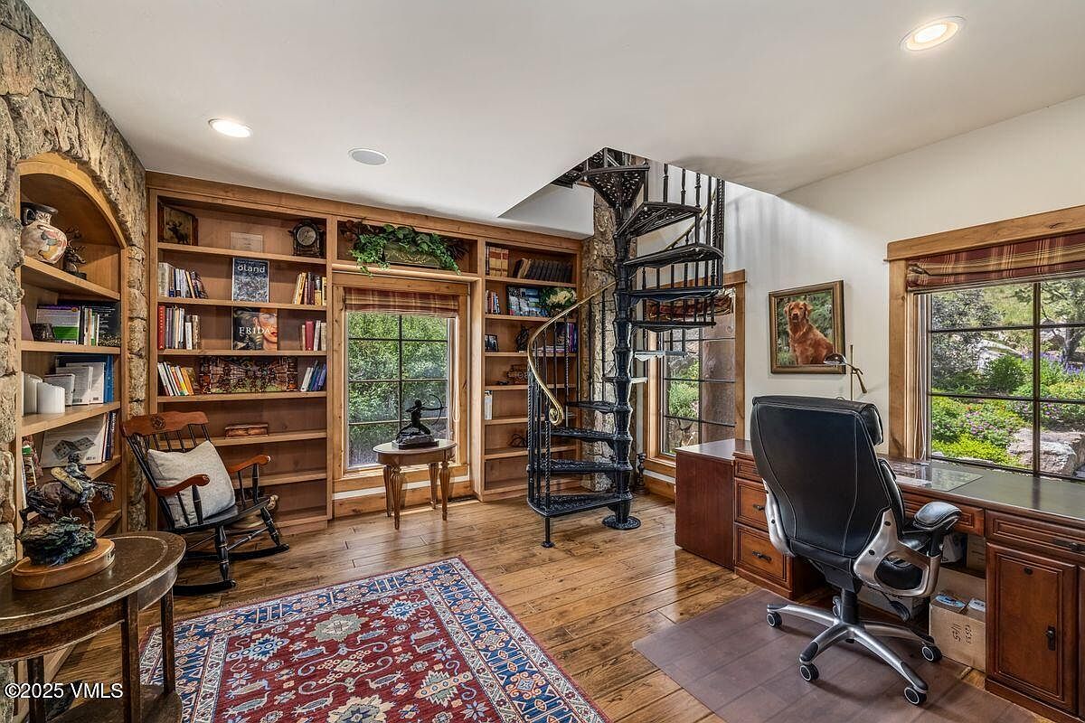 This is an inviting home office featuring custom built-in wooden bookshelves filled with books and decorative items. A spiral staircase adds a unique architectural element, while a large wooden desk with an executive chair provides a comfortable workspace. The room is well-lit with natural light from the windows, offering views of the outdoors.