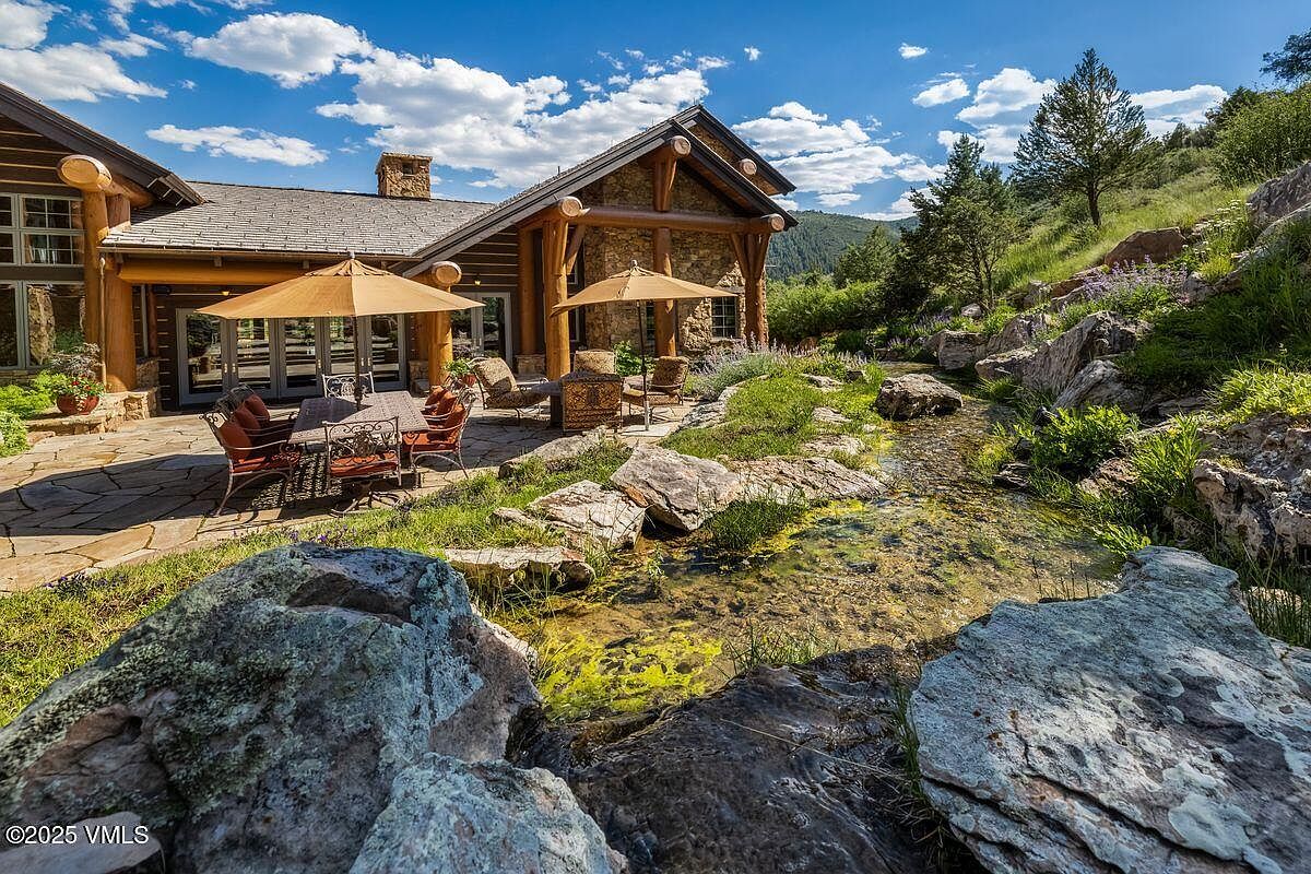 This exterior shot showcases a beautifully landscaped yard with a natural stream flowing through it. The property features a stone and wood house with outdoor seating areas shaded by umbrellas. The overall impression is one of tranquility and natural beauty, highlighting the property's outdoor living spaces.