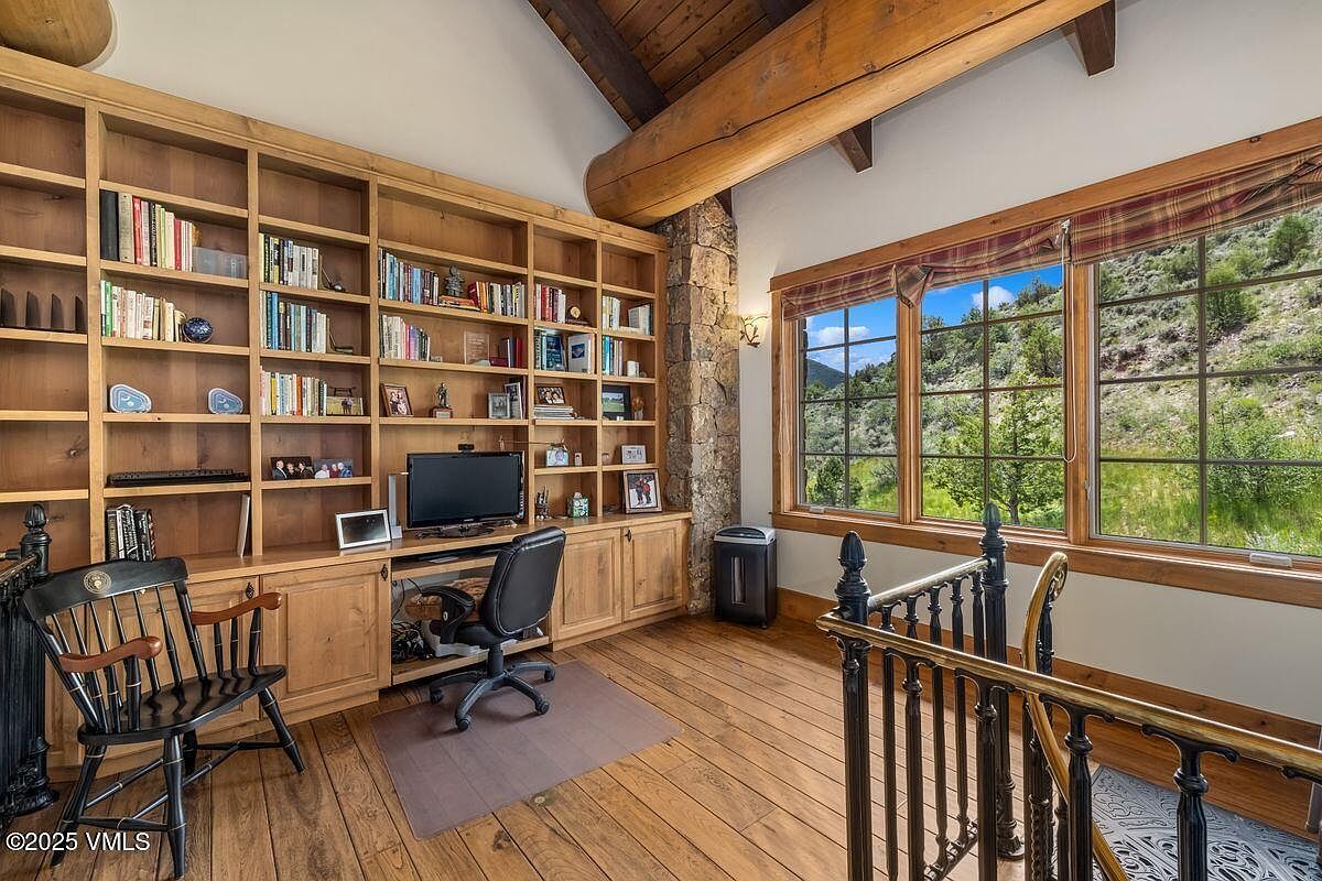 This is an interior shot of a home office or study, featuring a large built-in wooden bookcase filled with books and decorative items. A desk with a computer and office chair is positioned in front of the bookcase. A large window provides natural light and a view of the outdoors, while a stone pillar adds a rustic touch to the room's design.