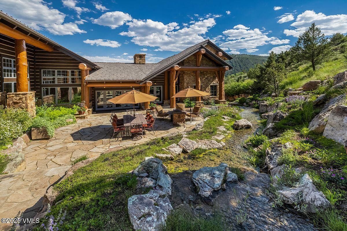 This exterior shot showcases a beautifully landscaped yard and garden area adjacent to a luxurious log cabin style home. A stone patio features outdoor dining and seating areas shaded by umbrellas, while a natural stream flows through the rocky terrain, adding to the serene ambiance. The scene is framed by lush greenery and mature trees, creating a picturesque and inviting outdoor space.