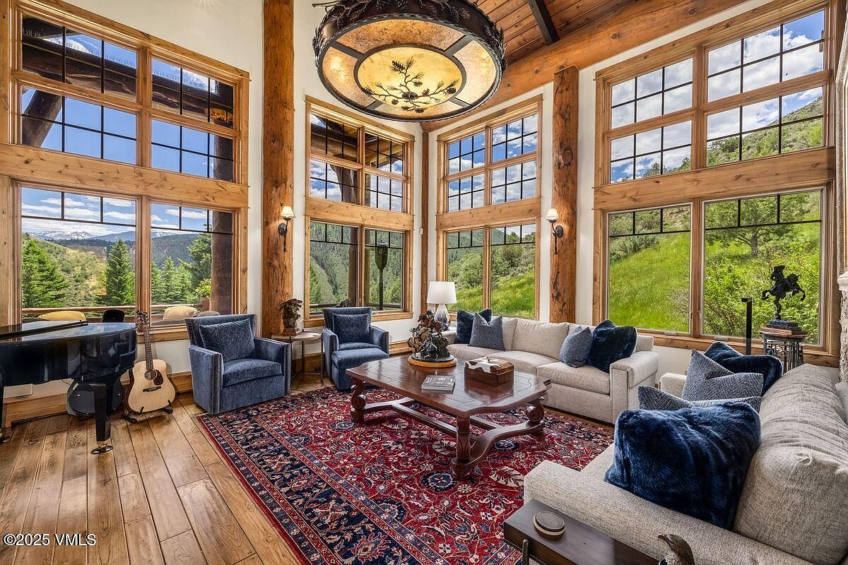 This is a spacious living room featuring large windows with views of the surrounding landscape. The room is furnished with comfortable sofas, armchairs, and a wooden coffee table, all arranged on a patterned rug. A grand piano and guitar are visible, adding a touch of elegance and musicality to the space.