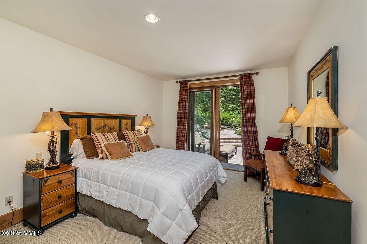 This is a cozy bedroom featuring a queen-sized bed with a white quilt and decorative pillows. The room has a warm and inviting atmosphere, with bedside tables, lamps, and a dresser providing ample storage. A sliding glass door leads to an outdoor patio, bringing natural light into the space.