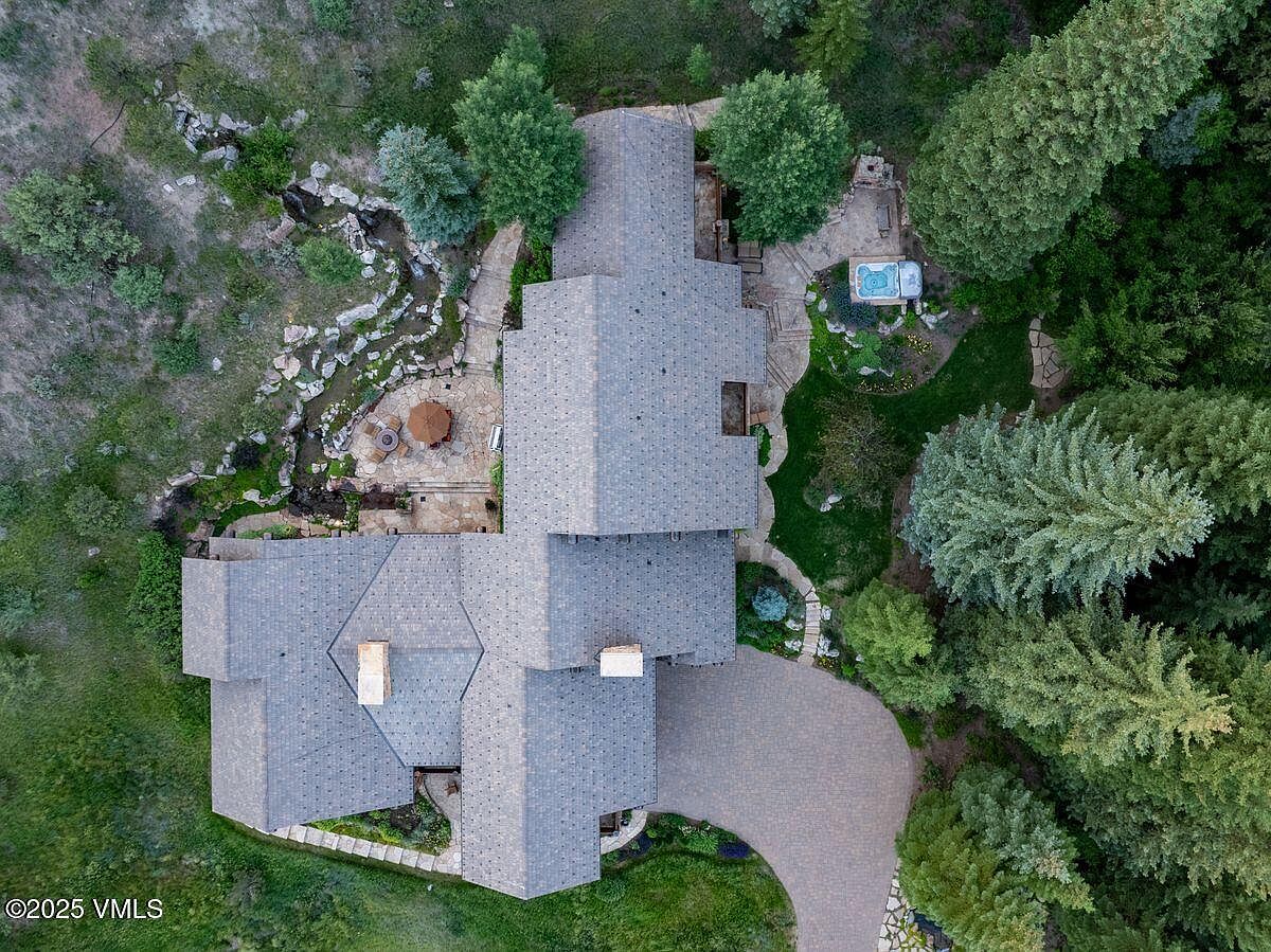 This aerial view showcases a luxurious home nestled amidst lush greenery and mature trees. The property features a meticulously landscaped yard with a stream, a patio area with an umbrella, and a hot tub, creating a serene outdoor oasis. A winding driveway leads to the house, which boasts a complex roofline and multiple chimneys.