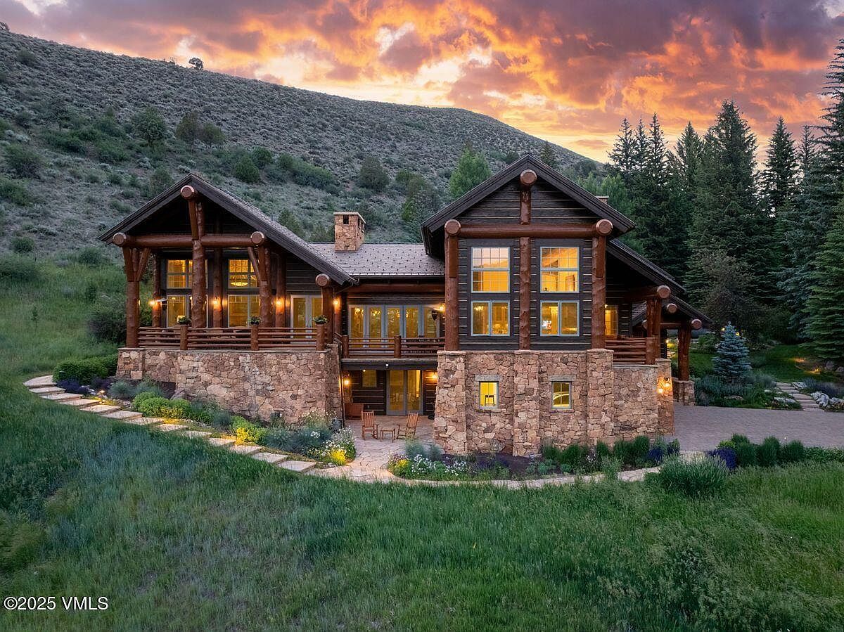 This is a stunning front view of a luxurious log cabin home. The house features a stone foundation, dark wood logs, and large windows that offer ample natural light. The surrounding landscape includes a well-manicured lawn, stone pathways, and a backdrop of lush greenery and a mountain, creating a serene and private setting.