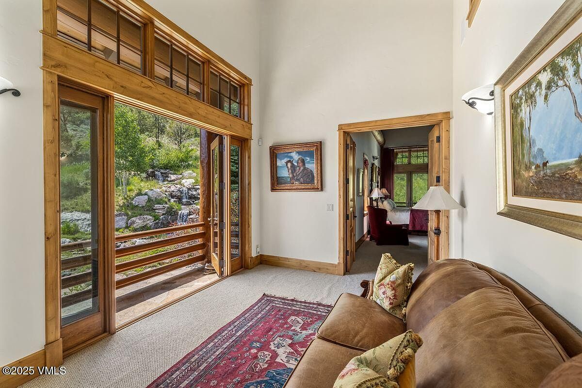 This interior shot showcases a cozy living room with a rustic-chic aesthetic. A comfortable brown leather sofa sits atop a patterned rug, with a view to the outside through large wooden-framed windows and doors that lead to a balcony overlooking a waterfall. The room also features artwork and a doorway leading to another room, suggesting a spacious and inviting home.
