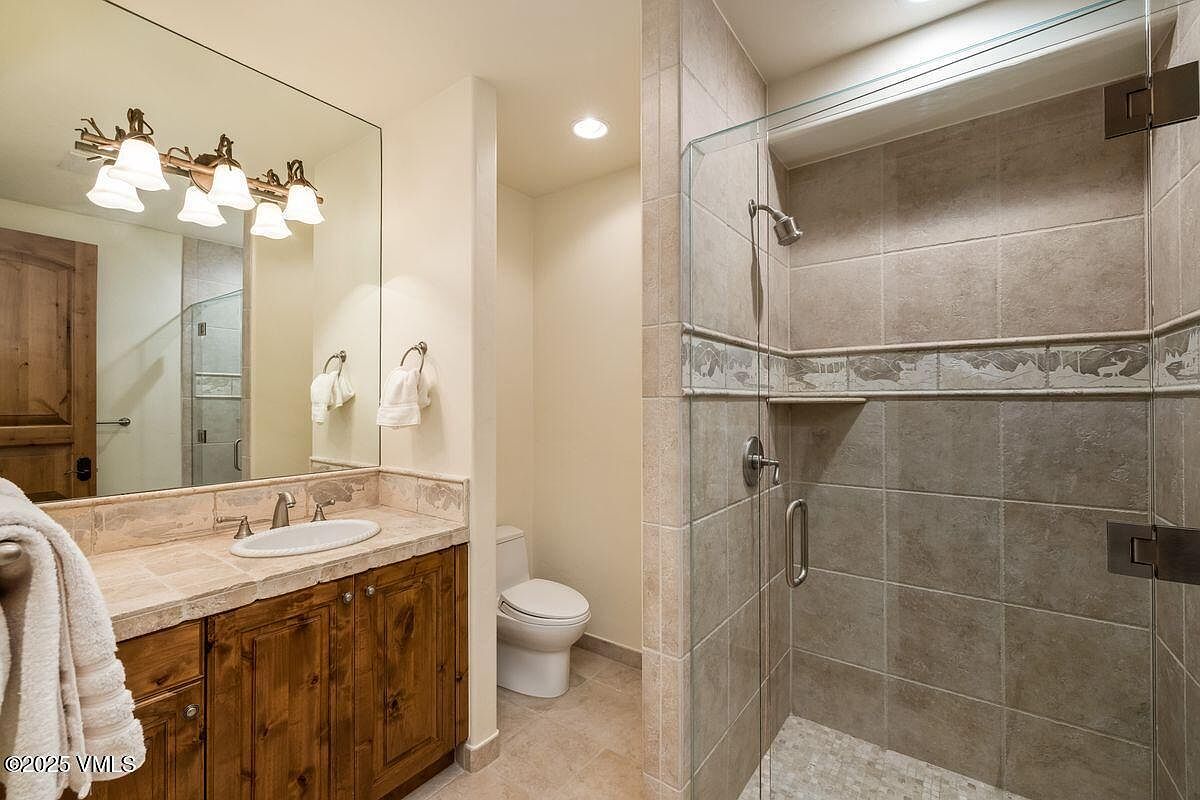 This is a well-lit bathroom featuring a vanity with a wooden cabinet and light-colored countertop, a toilet, and a glass-enclosed shower with tiled walls. The mirror reflects the vanity area, creating a sense of spaciousness. The overall style is clean and functional, suitable for a guest bathroom.