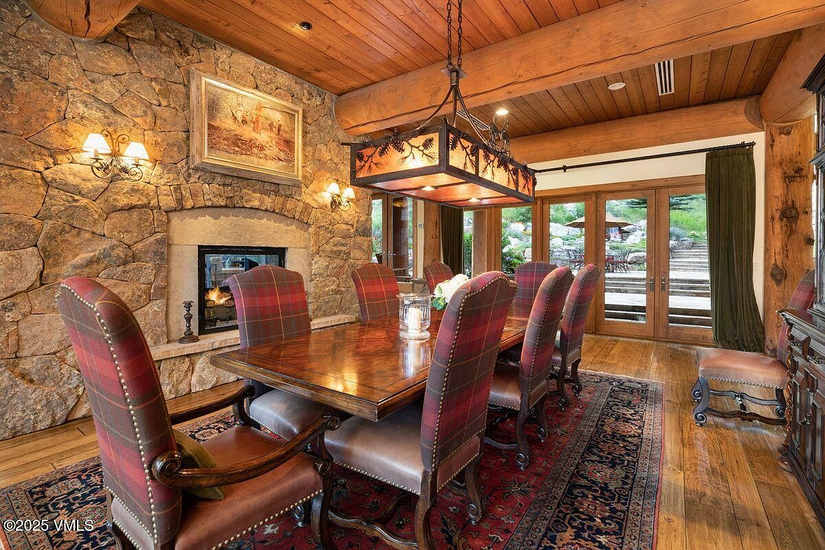 This dining room features a rustic yet elegant design with a stone wall, a wooden ceiling with exposed beams, and a rectangular wooden dining table surrounded by upholstered chairs with plaid backs. A decorative chandelier hangs above the table, and French doors lead to an outdoor area, creating a warm and inviting atmosphere perfect for family gatherings or formal dinners.