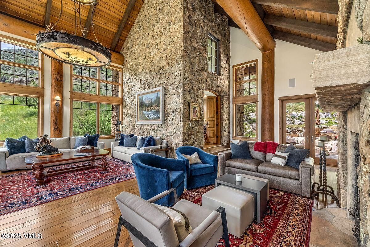 This is an interior shot of a luxurious living room featuring a high vaulted ceiling with wooden beams and a stone accent wall. The room is furnished with comfortable sofas, armchairs, and a coffee table, all arranged on a patterned rug over hardwood floors. Large windows provide ample natural light and views of the surrounding landscape, creating a warm and inviting atmosphere.