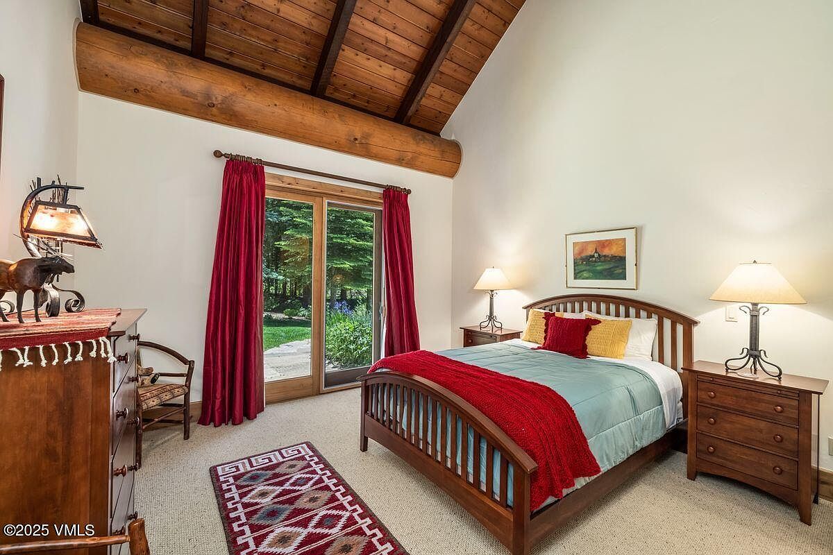 This is a cozy bedroom featuring a wooden bed frame with a red throw blanket and decorative pillows. The room has a rustic charm with exposed wooden beams on the ceiling and a view to the outside through a glass door with red curtains. A wooden dresser and nightstands with lamps add to the warm and inviting atmosphere.