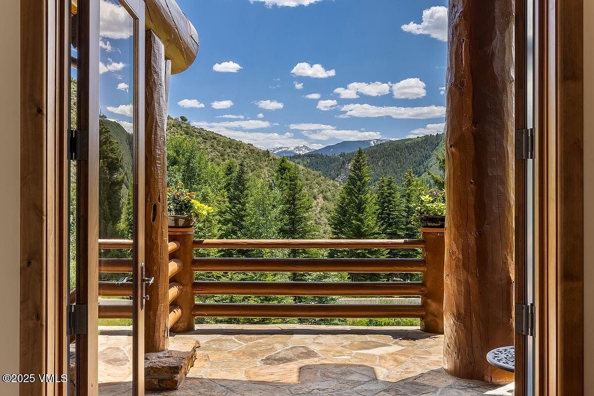 This image showcases a rustic patio or balcony with a stunning mountain view. The space features a stone floor and a wooden railing, complemented by substantial log pillars. The open doorway frames a picturesque scene of lush green trees and distant snow-capped mountains under a bright blue sky with scattered clouds, creating a serene and inviting atmosphere.