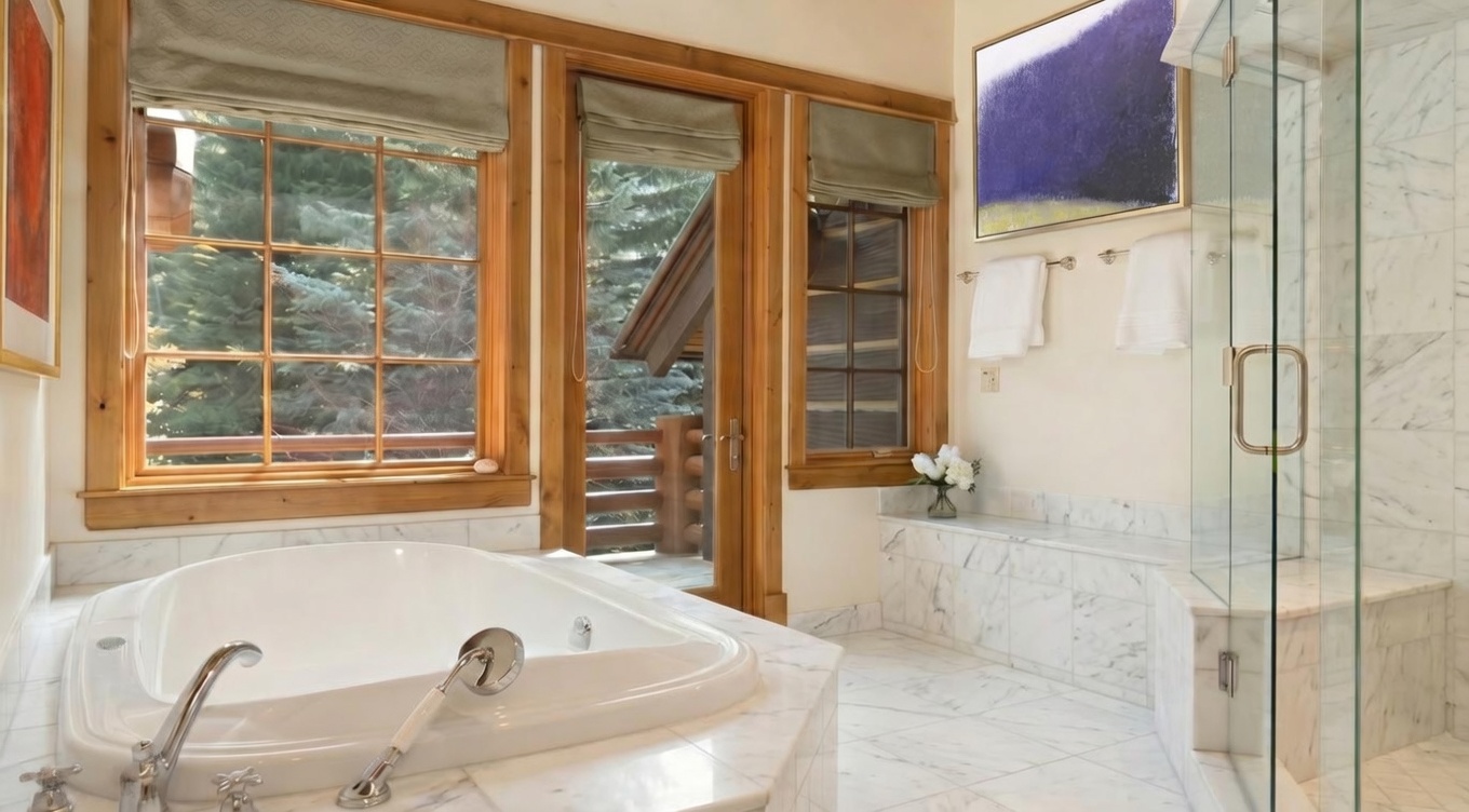 This is a luxurious primary bathroom featuring a large soaking tub with chrome fixtures, set against a backdrop of natural wood-framed windows offering a view of the outdoors. The bathroom is finished with travertine tile, a glass-enclosed shower, and a built-in bench, creating a spa-like atmosphere.