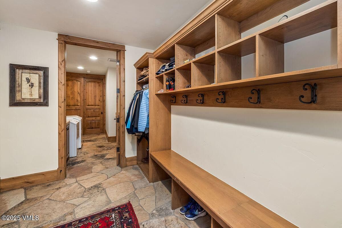 This interior shot showcases a hallway with a mudroom area. The space features custom wooden built-ins with shelving, hooks, and a bench, providing ample storage. The flooring is a natural stone tile, and a framed artwork hangs on the wall, adding a touch of elegance to the functional space.
