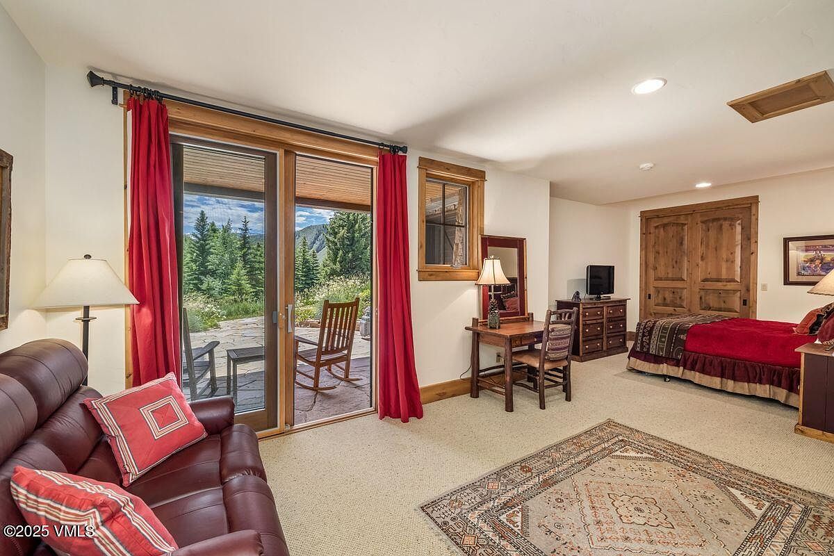 This is a cozy guest bedroom featuring a comfortable leather sofa with red pillows, a wooden desk and chair, and a bed with red bedding. A sliding glass door leads to an outdoor patio, offering a view of the surrounding greenery. The room is carpeted and has a warm, inviting atmosphere.