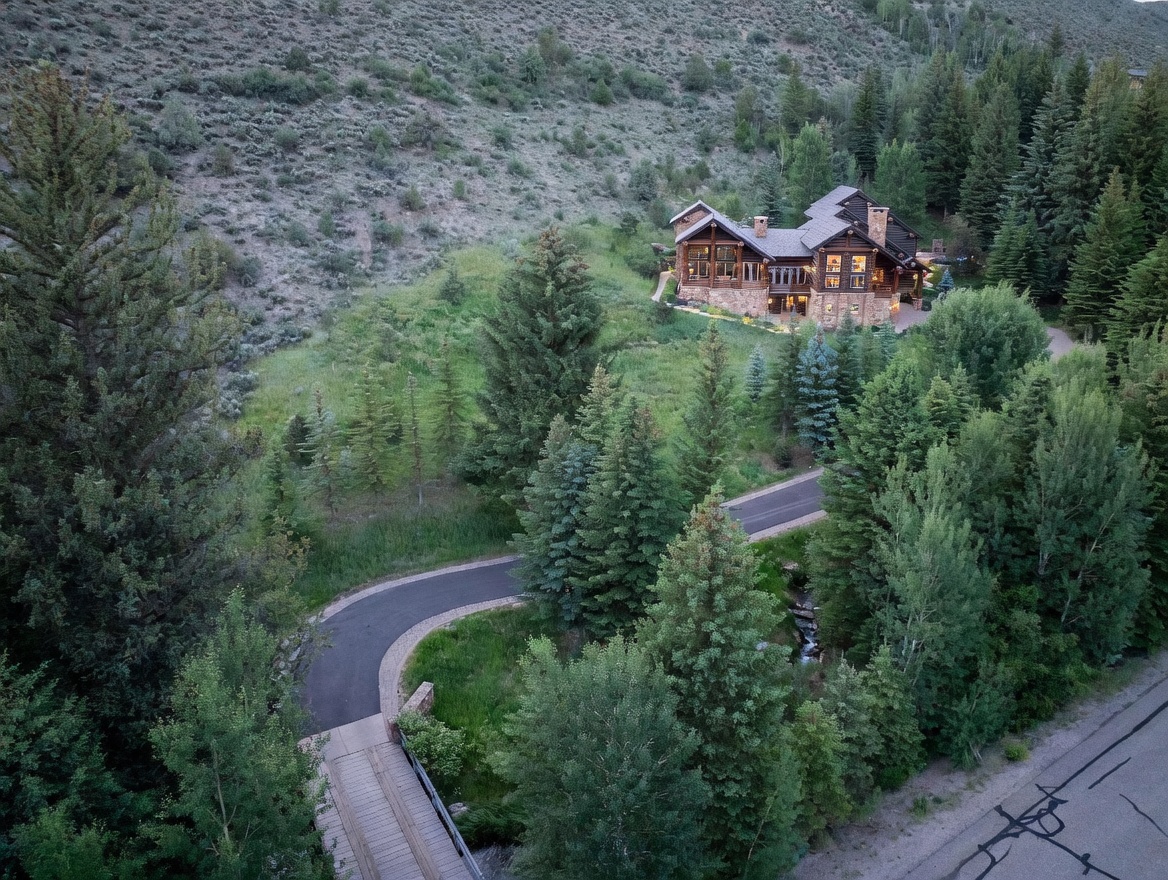 This aerial view showcases a luxurious mountain home nestled among lush greenery and mature trees. A winding driveway leads to the residence, which features a stone base and wood construction, blending seamlessly with the natural surroundings. The overall impression is one of secluded elegance and natural beauty.
