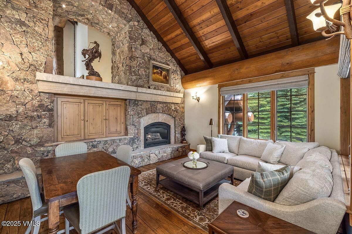This inviting living room features a stone fireplace with a wooden mantel and built-in cabinetry, creating a cozy focal point. A comfortable sectional sofa and a leather ottoman offer ample seating, while a wooden dining table with upholstered chairs provides a space for meals or games. The room is bathed in natural light from a large window, showcasing a blend of rustic charm and modern comfort.