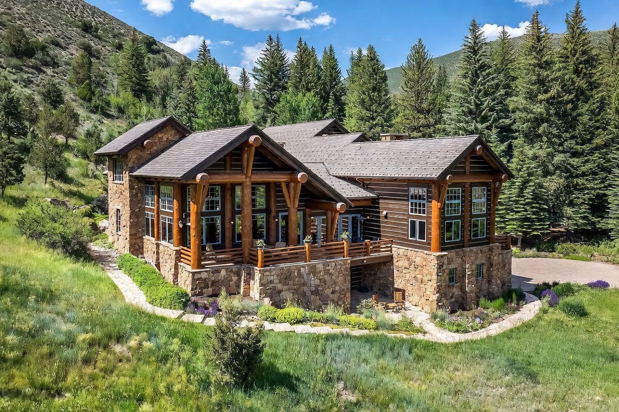 This is a front exterior view of a luxurious mountain home. The house features a combination of stone and wood construction, with large windows and a prominent covered porch area. The surrounding landscape includes lush greenery and mature trees, creating a serene and private setting.
