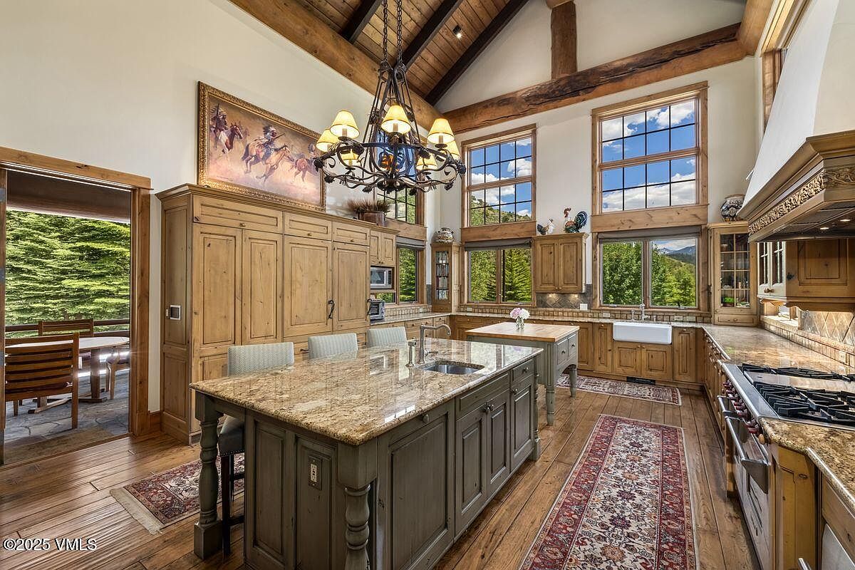 This is a spacious kitchen with a rustic yet luxurious design. It features wooden cabinetry, granite countertops, a large central island with seating, and high ceilings with exposed wooden beams. Large windows provide ample natural light and views of the surrounding landscape, enhancing the kitchen's inviting atmosphere.