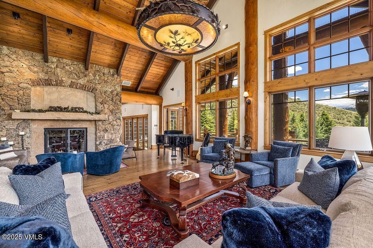 This is an interior shot of a luxurious living room featuring a stone fireplace, wood-beamed ceilings, and large windows offering scenic views. The room is furnished with comfortable sofas, armchairs, and a grand piano, creating an inviting and elegant atmosphere. A large, ornate chandelier hangs from the ceiling, adding to the room's grandeur.