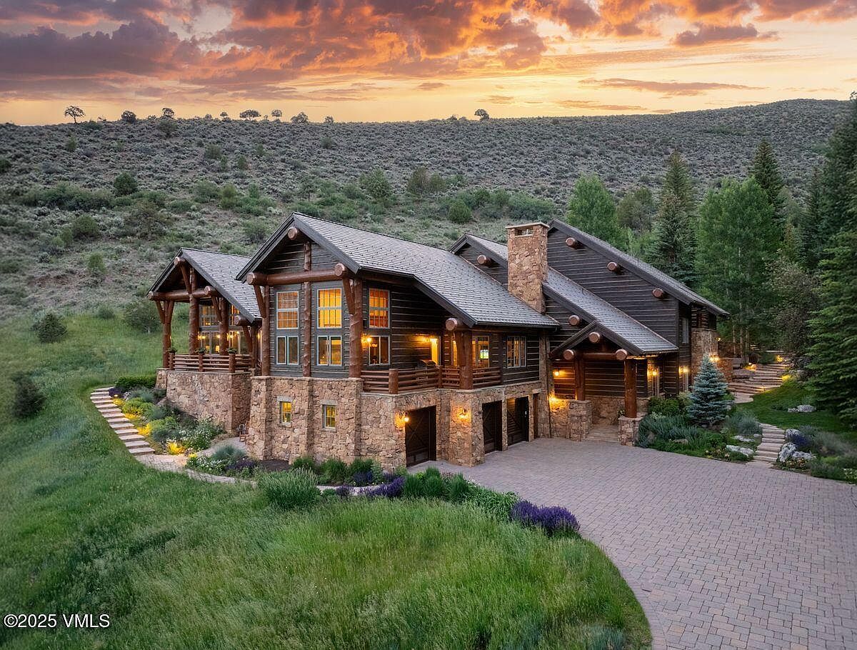 This is a stunning front view of a luxurious log cabin home. The exterior features a combination of stone and wood, with a well-manicured lawn and landscaping. A paved driveway leads to the garage, and the home is nestled against a backdrop of rolling hills and a vibrant sunset sky, creating a picturesque and inviting scene.