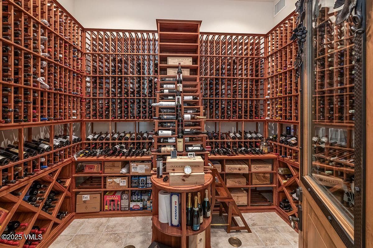 This is a well-stocked wine cellar featuring extensive wooden racking that spans floor to ceiling on multiple walls. A central wooden structure with shelves and a small ladder provides access to higher shelves, and various wine bottles and wooden cases are displayed throughout. The room is illuminated with warm lighting, creating an inviting atmosphere for wine enthusiasts.