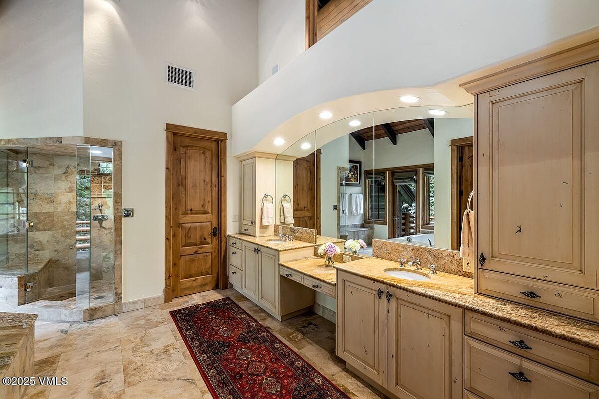This is a primary bathroom featuring a double vanity with light wood cabinetry and granite countertops. A large, arched mirror with integrated lighting spans the length of the vanity. A glass-enclosed shower is visible to the left, and a decorative rug adds warmth to the tiled floor. The room has high ceilings, enhancing the sense of space.
