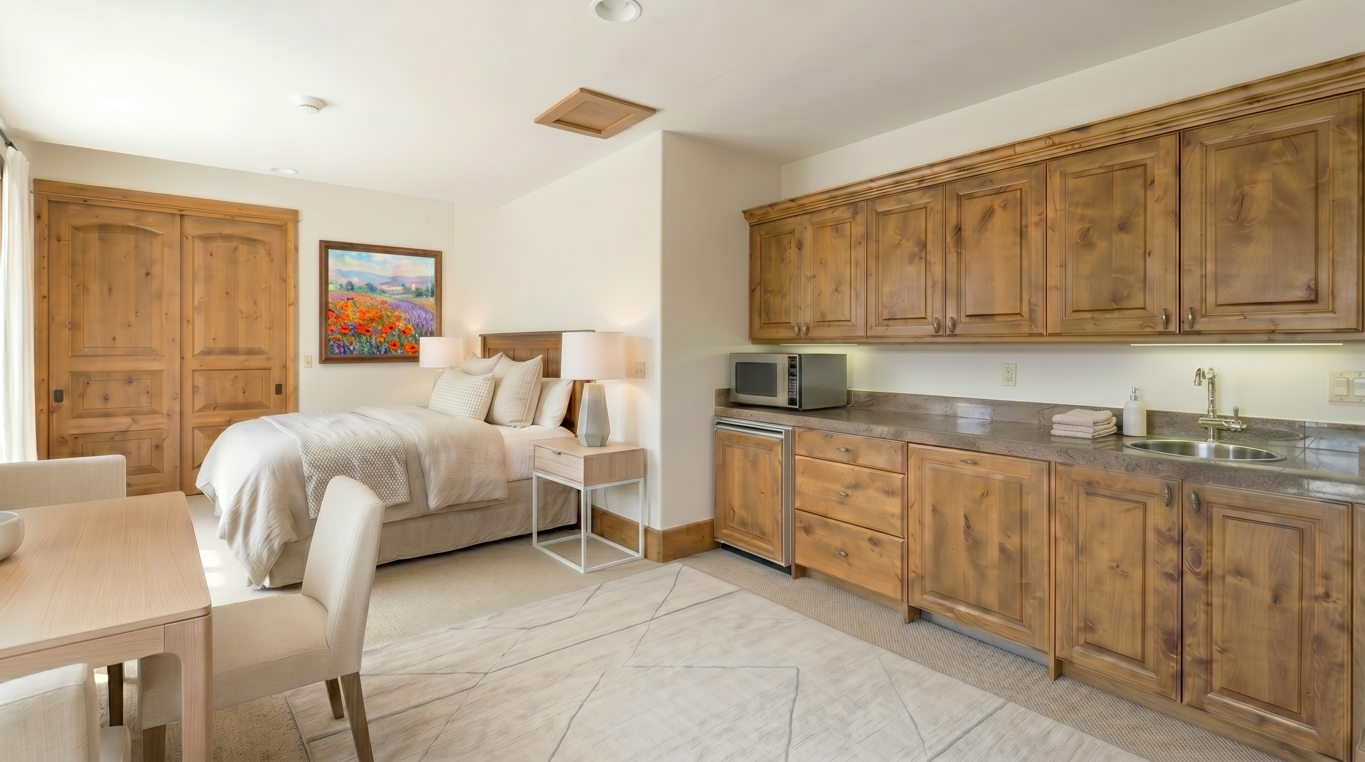 This is a cozy guest bedroom featuring a comfortable bed with red bedding, a wooden nightstand with a lamp, and a built-in kitchenette area with wooden cabinets, a microwave, and a sink. The room is carpeted and includes a decorative rug, adding warmth and character. The overall impression is inviting and functional, suitable for a comfortable guest stay.
