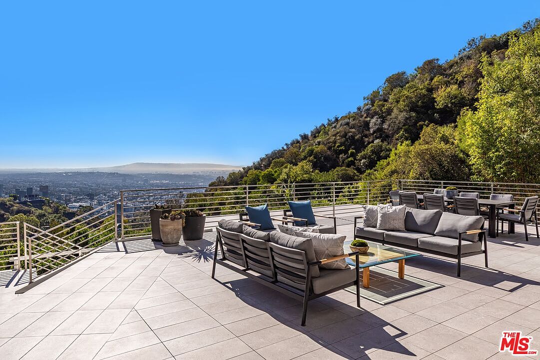 This expansive outdoor terrace offers a panoramic view of the city skyline set against a backdrop of lush, tree-covered hills. The space is furnished with modern, dark-framed lounge seating and a dining set, all arranged on large, neutral-toned stone tiles. A sleek metal railing borders the deck, emphasizing the open-air, elevated perspective of this luxurious hillside property.