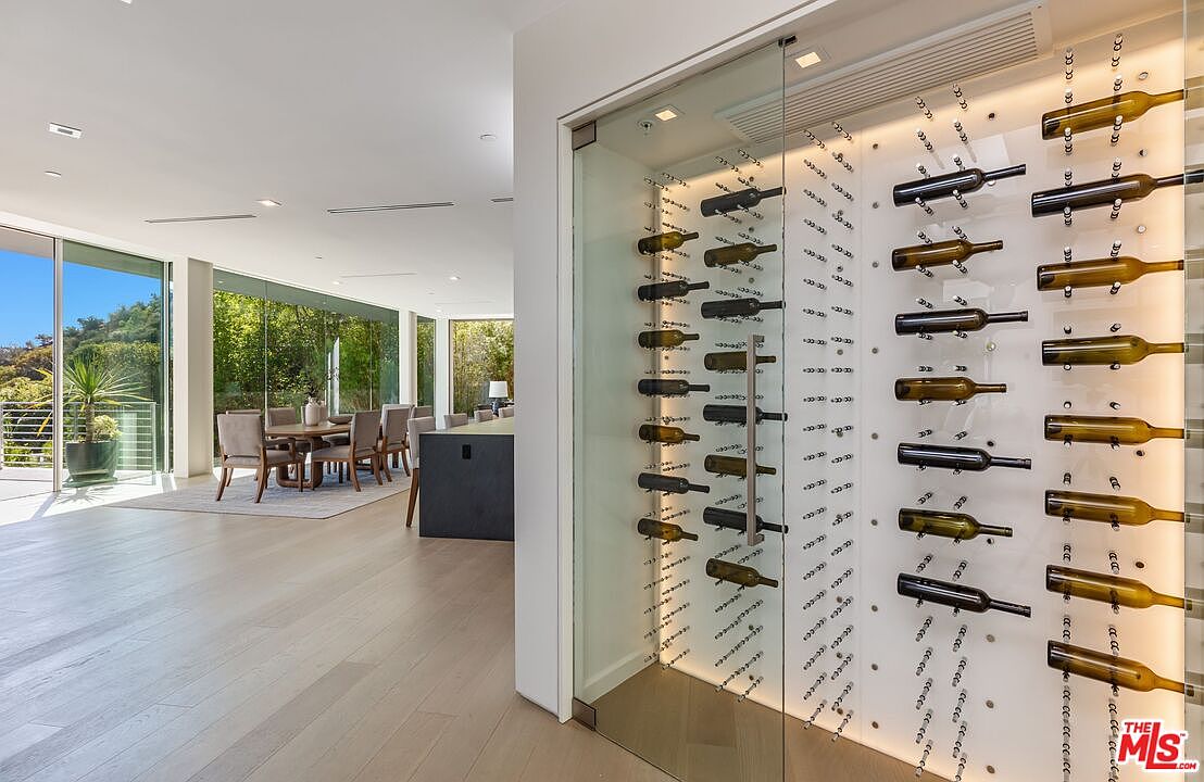 This image showcases a sophisticated, climate-controlled glass wine cellar integrated into a modern open-concept living space. The cellar features a minimalist design with horizontal bottle racks mounted against a backlit panel, creating a striking visual display. Beyond the cellar, the view extends into a bright dining area with large windows that provide a seamless connection to the lush outdoor greenery.