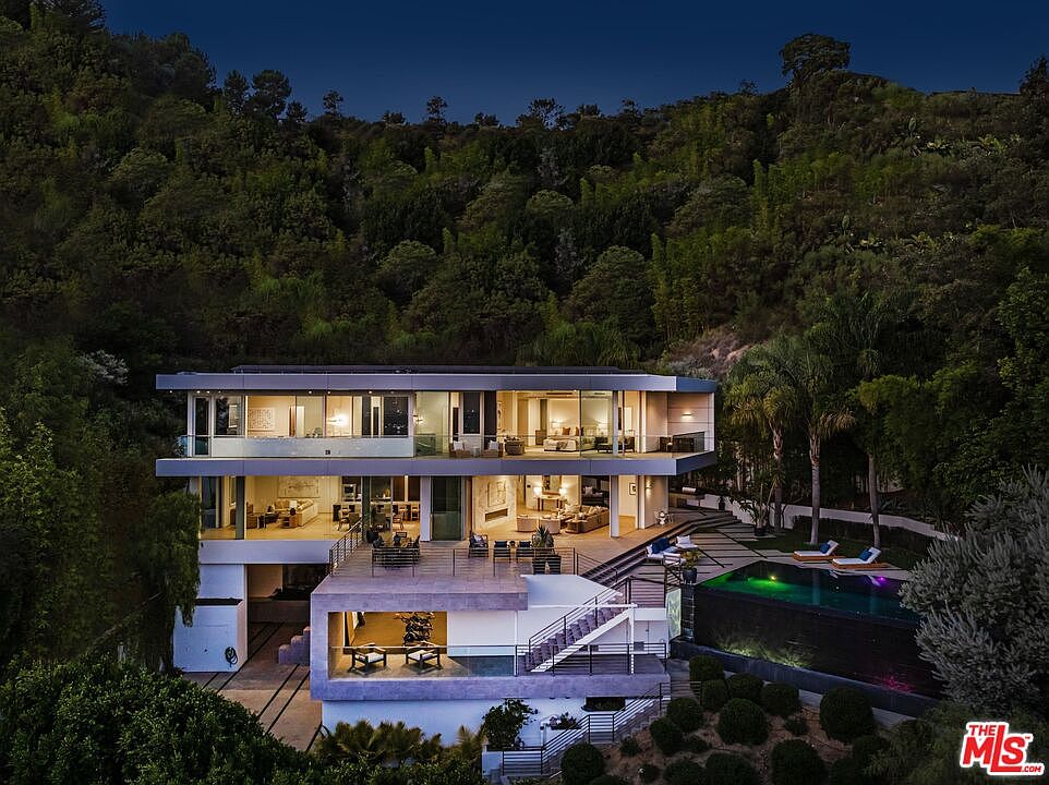 This striking modern residence is captured from an elevated perspective at dusk, showcasing its multi-level architectural design nestled against a lush, forested hillside. The home features expansive glass walls, multiple terraces for outdoor living, and a sleek infinity-edge pool that glows with integrated lighting. The composition highlights the seamless integration of contemporary luxury living with the natural landscape, emphasizing the property's dramatic scale and sophisticated aesthetic.