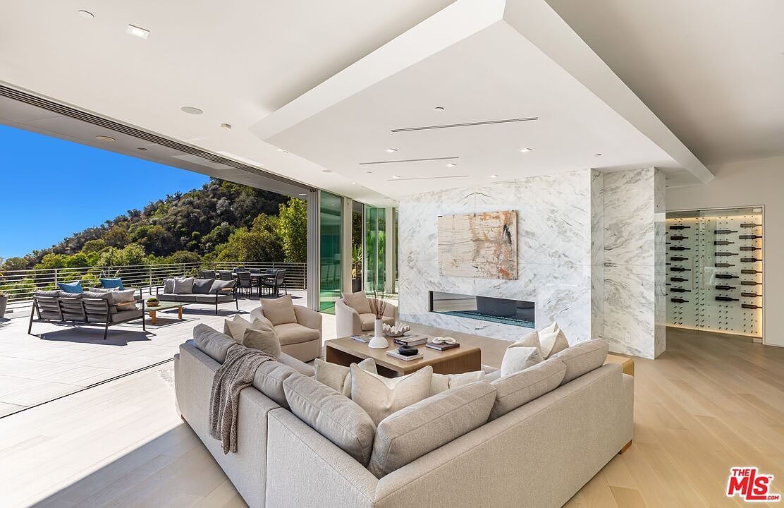 This expansive, open-concept living room features a neutral color palette and seamless indoor-outdoor flow through large sliding glass doors that open to a spacious deck. A central stone-clad wall houses a linear fireplace, while a glass-enclosed wine display adds a sophisticated touch to the side. The room is furnished with a large sectional sofa and comfortable armchairs, creating an inviting space that overlooks a lush, hilly landscape.
