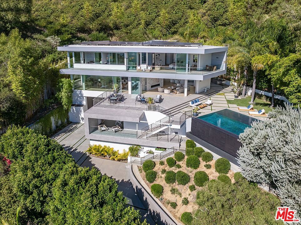 This high-angle aerial view captures a modern, multi-level luxury residence nestled into a lush, hillside landscape. The home features clean, horizontal lines, extensive glass walls, and multiple expansive terraces that provide seamless indoor-outdoor living. A sleek, dark-tiled infinity pool sits adjacent to a paved patio area, surrounded by manicured landscaping and mature trees, creating a sophisticated and private retreat.