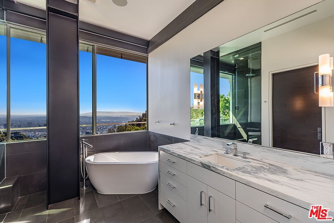 This modern primary bathroom features a sleek, freestanding soaking tub positioned next to large windows that offer an expansive, elevated view of the city skyline. A long vanity with a stone countertop and integrated sink provides ample storage, complemented by a large wall-mounted mirror and contemporary light fixtures. The space is defined by its clean lines, dark tile flooring, and a sophisticated, minimalist aesthetic.