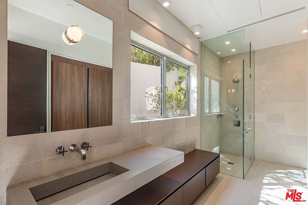 This modern bathroom features a sleek, integrated stone sink vanity with a dark wood cabinet base and a large, wall-mounted mirror. To the right, a glass-enclosed walk-in shower is finished with light-toned stone tiles, complemented by a window that brings in natural light. The space is designed with a minimalist aesthetic, emphasizing clean lines and high-quality materials.