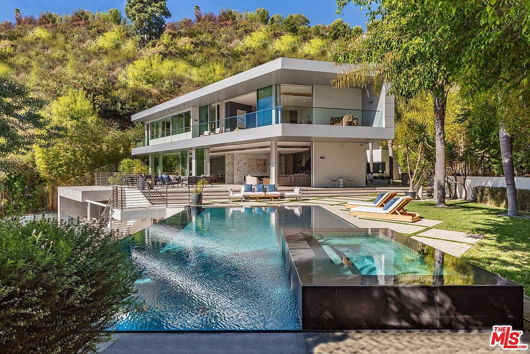 This modern luxury residence features a striking architectural design with clean lines, expansive glass walls, and a multi-level structure nestled against a lush, hillside backdrop. The rear exterior showcases a seamless transition between the indoor living spaces and a spacious patio, which overlooks a sleek infinity-edge pool and integrated spa. The composition emphasizes a sophisticated, resort-like atmosphere, highlighting the property's integration with its natural environment.